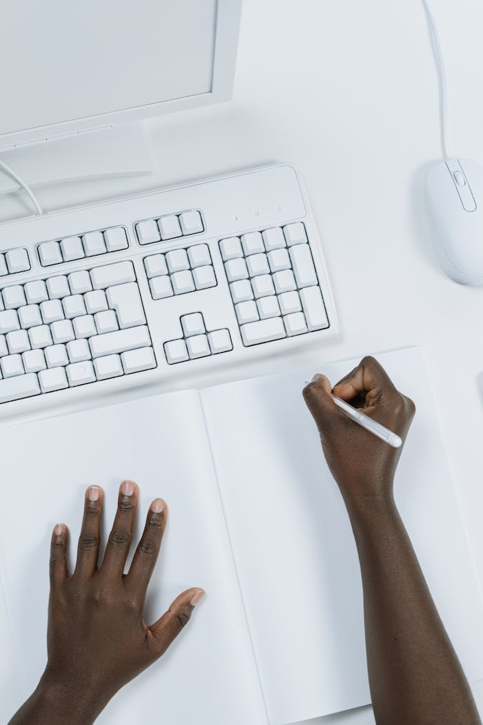 Overhead view of hands writing in a notebook at a clean, minimalist workspace with a keyboard and mouse.